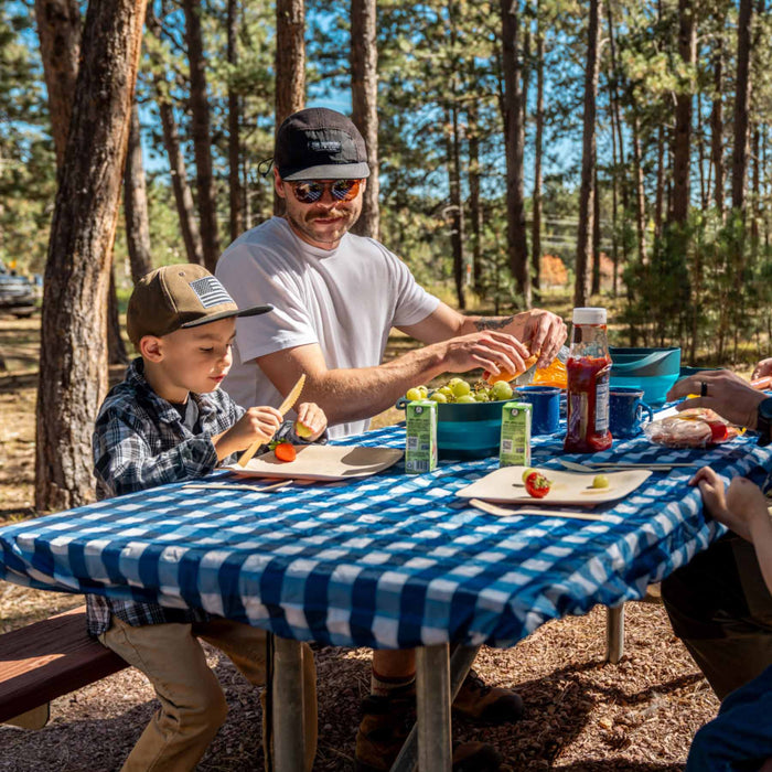 A father and child eating fruits at a picnic table covered in the Adventure Ready Pincnic Tablecloth. The child is using the bamboo utensil from the Adventure Ready Bamboo Dining Set and has fruits on his bamboo plate.